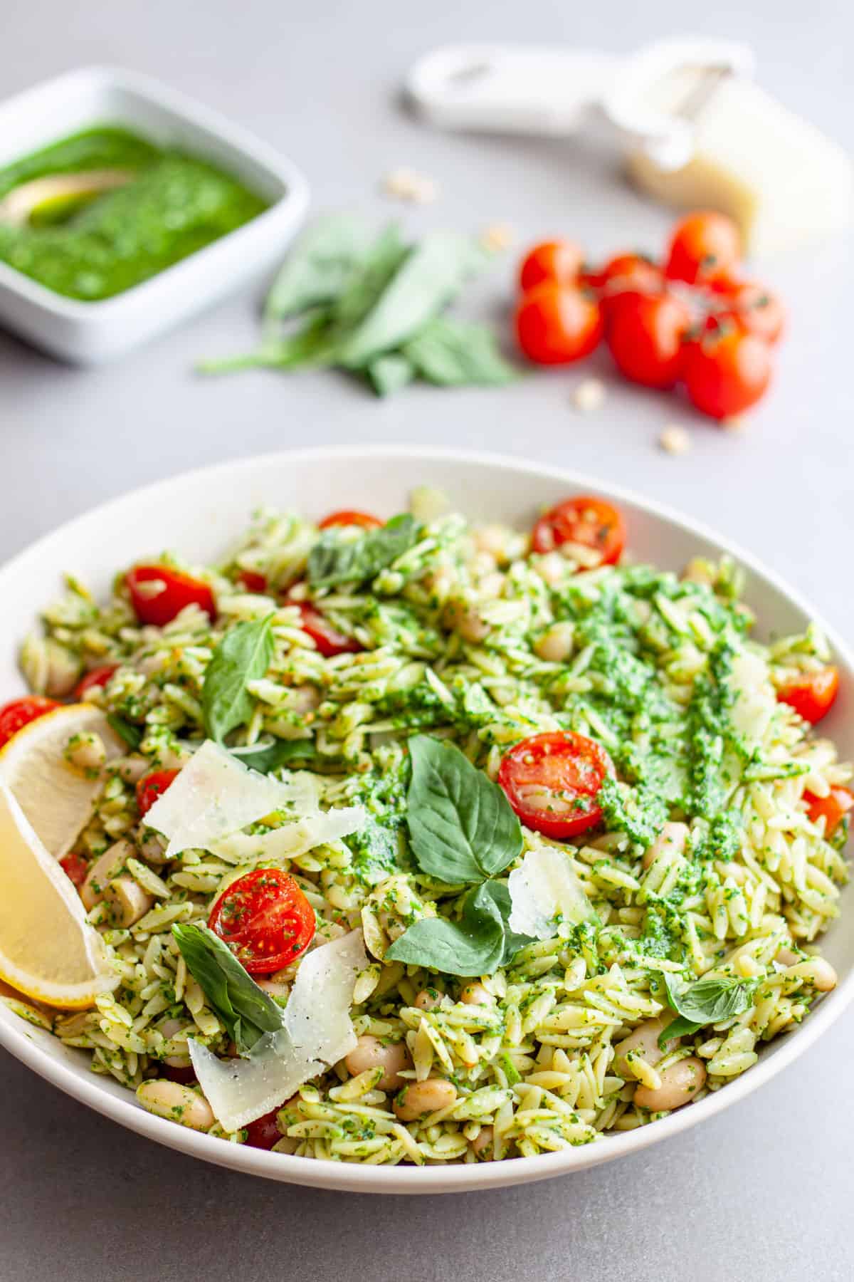 A large white serving bowl of orzo with pesto and tomatoes on a gray table.