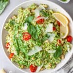 A white serving bowl of pesto pasta with chopped tomatoes, basil leaves and shaved parmesan on top.