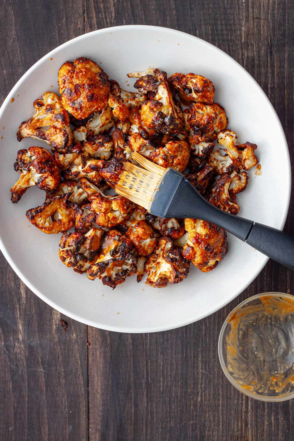 A white bowl with roasted cauliflower getting brushed with a miso sauce.
