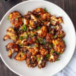 A white bowl of roasted miso cauliflower topped with thinly sliced scallions on a dark table.