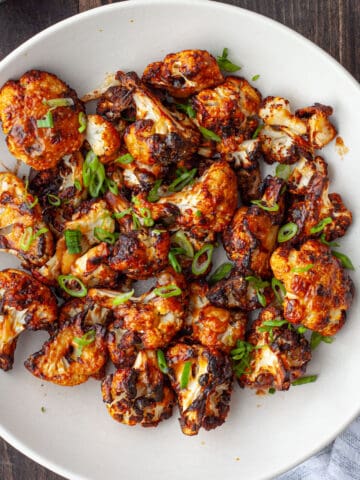 A white bowl of roasted miso cauliflower topped with thinly sliced scallions on a dark table.