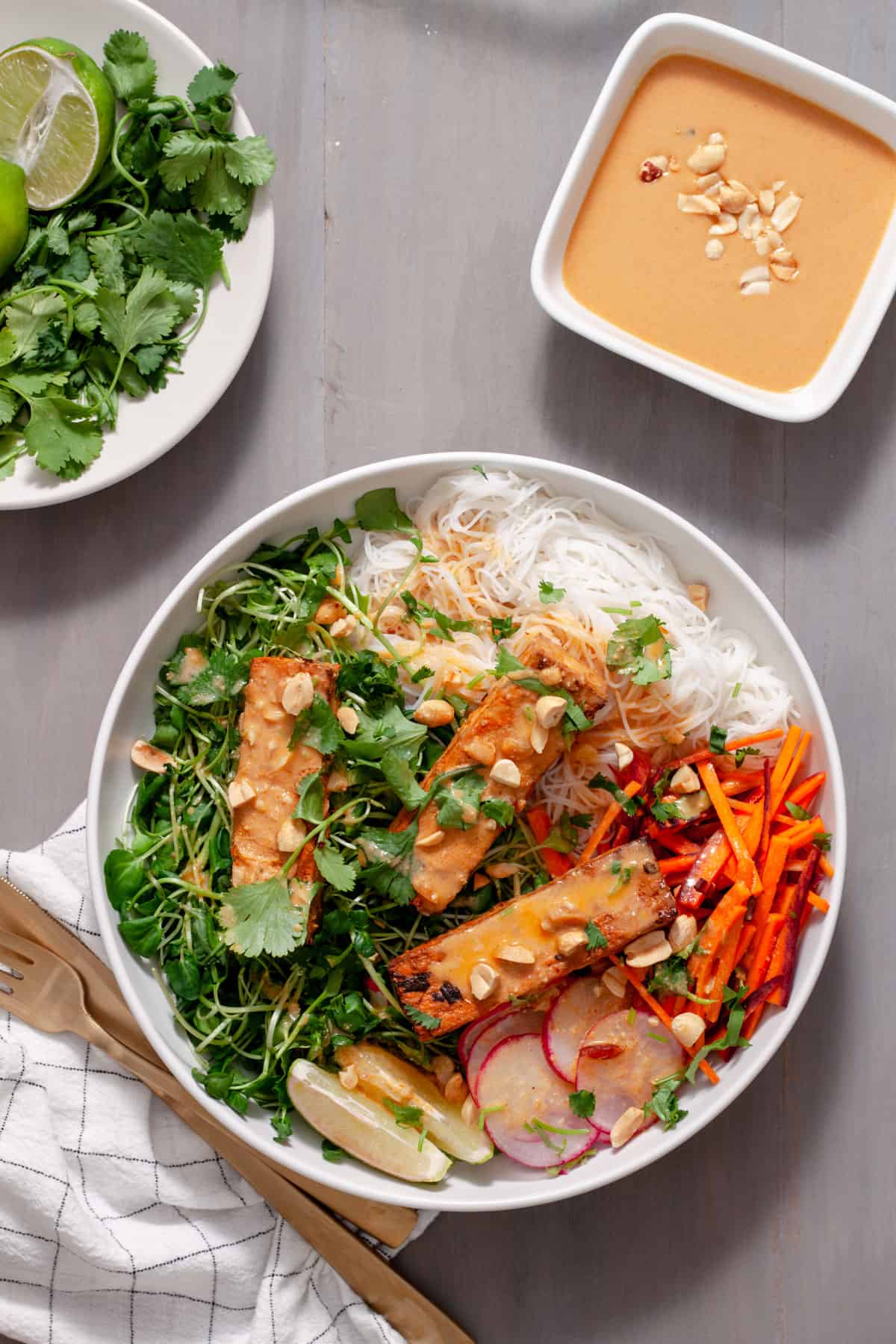 A white bowl with rice noodles, watercress, carrots and crispy tofu topped with peanut sauce.