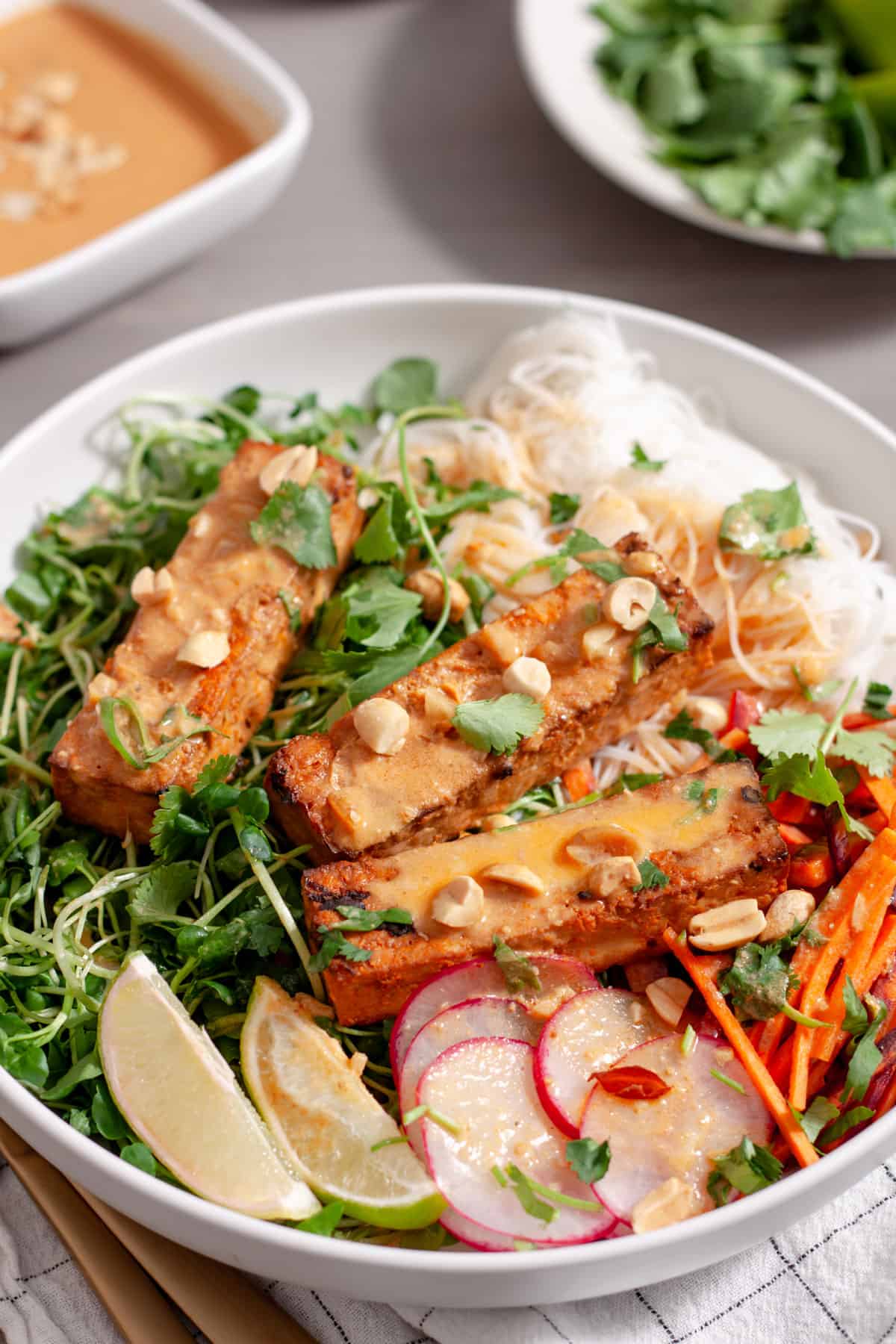 A white bowl with a summer roll salad topped with crispy peanut tofu, herbs, peanuts and peanut sauce.