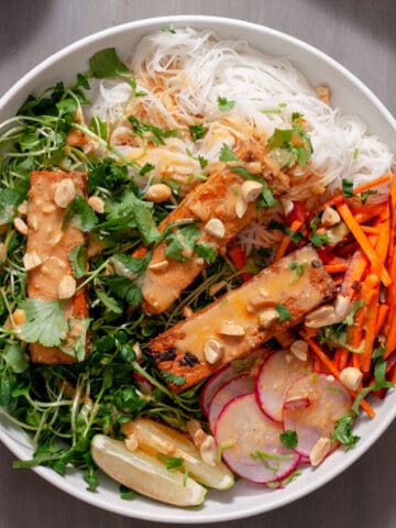 A white bowl with a summer roll salad topped with crispy peanut tofu, herbs, peanuts and peanut sauce.