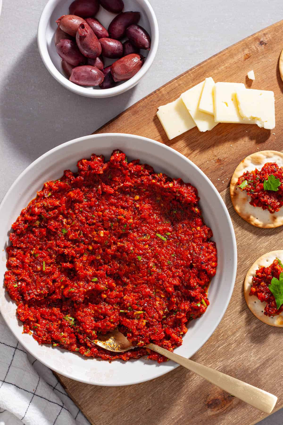 A bowl of sun-dried tomato spread with olives to the side and crackers on a cutting board.