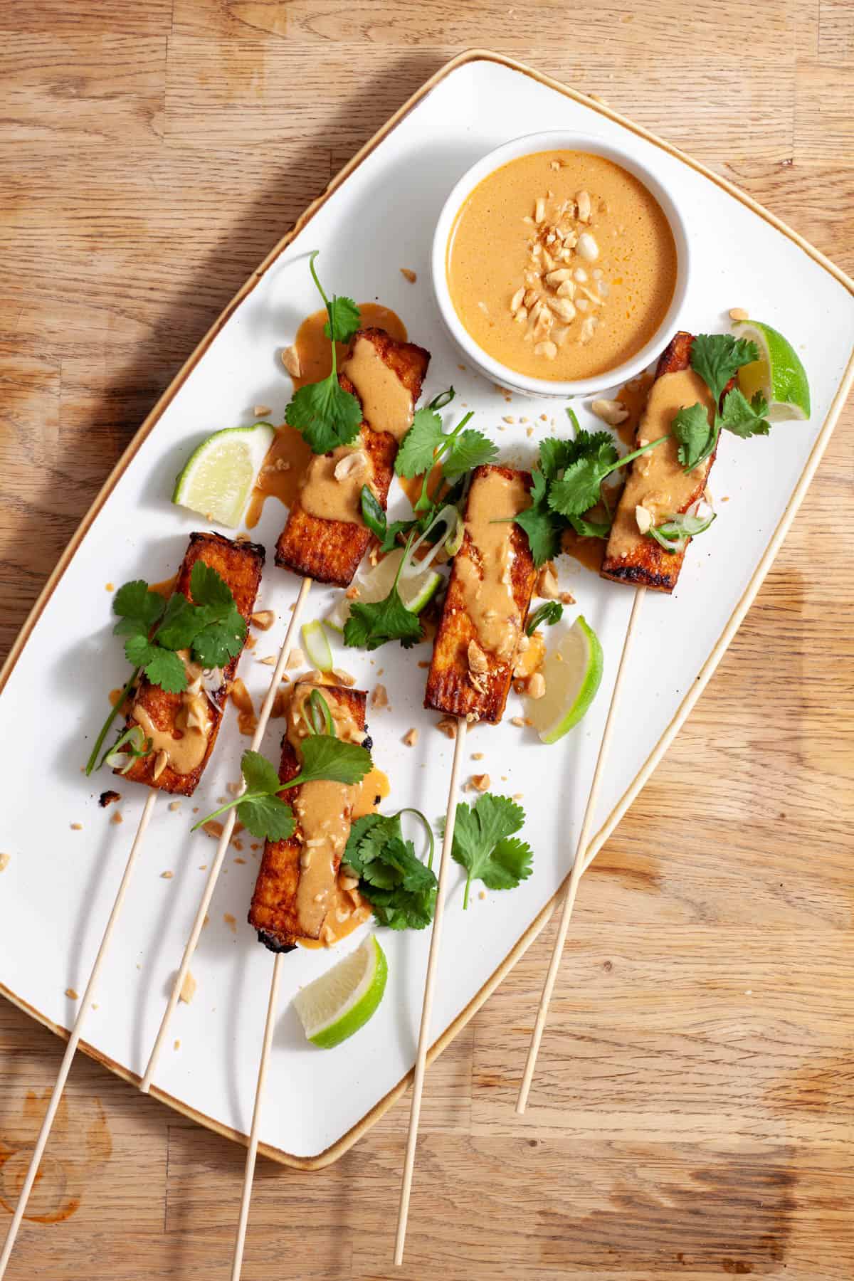 A platter of tofu satay topped with cilantro, peanuts and a peanut sauce on a butcherblock counter.