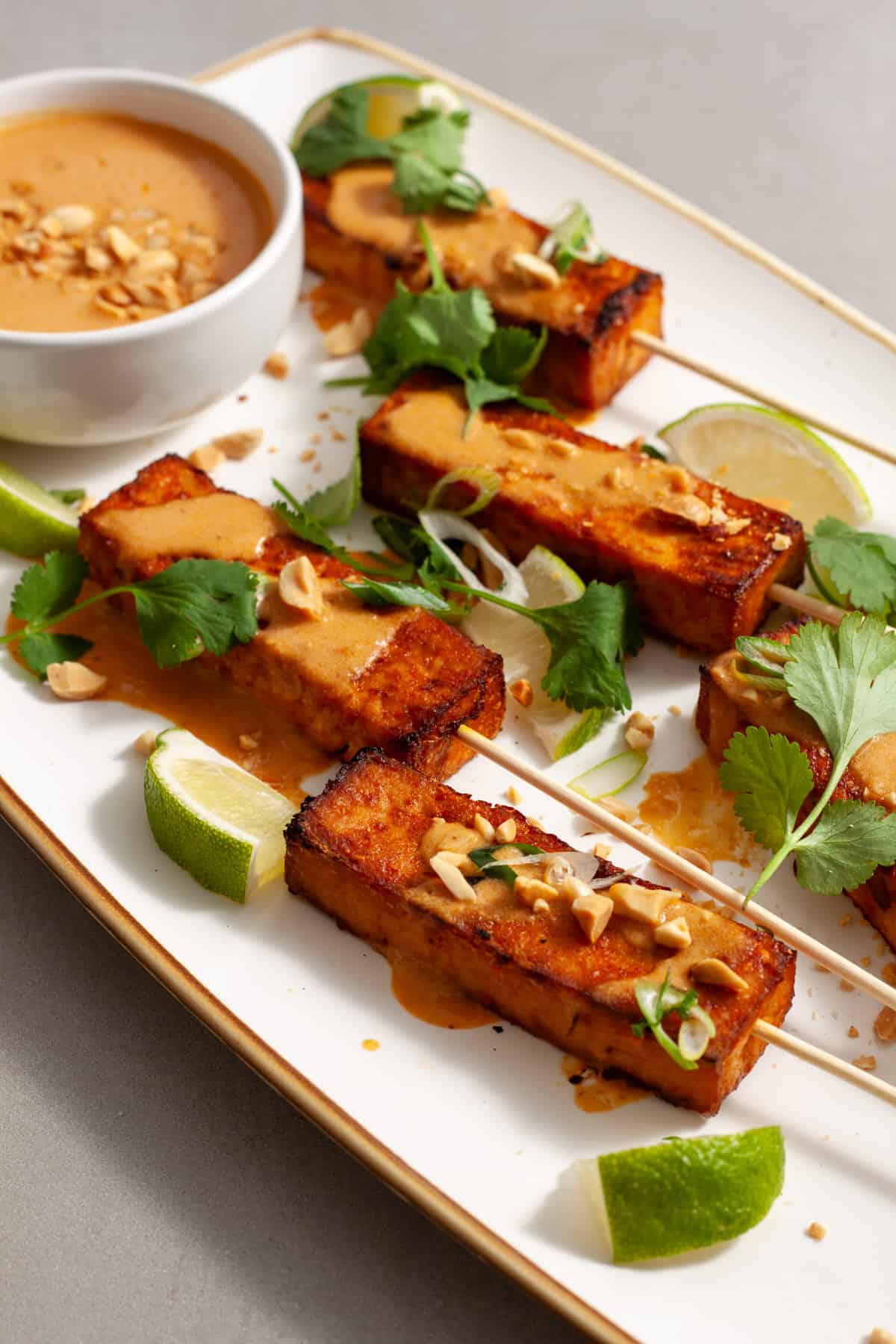 A white platter of tofu satay topped with peanut sauce, herbs and chopped peanuts.