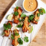 A platter of tofu satay topped with cilantro, peanuts and a peanut sauce on a butcherblock counter.