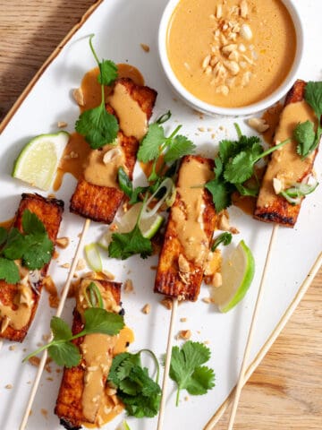 A platter of tofu satay topped with cilantro, peanuts and a peanut sauce on a butcherblock counter.