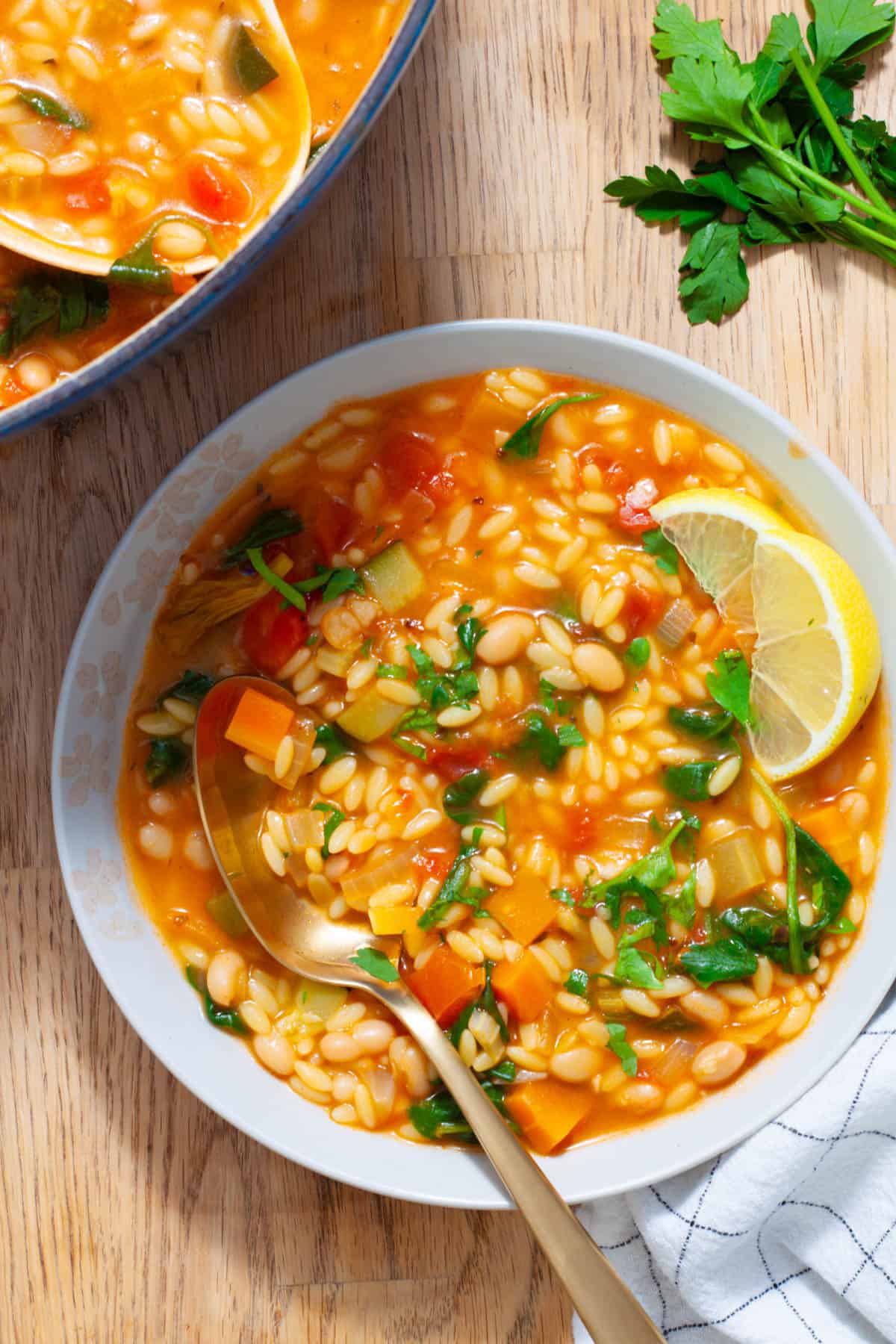 A bowl of orzo vegetable soup with lemon wedges to the side and topped with parsley.