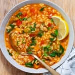 A white bowl of vegetable orzo soup topped with chopped parsley and lemon wedges on the side.