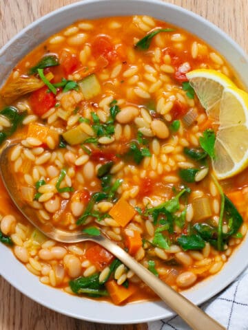 A white bowl of vegetable orzo soup topped with chopped parsley and lemon wedges on the side.