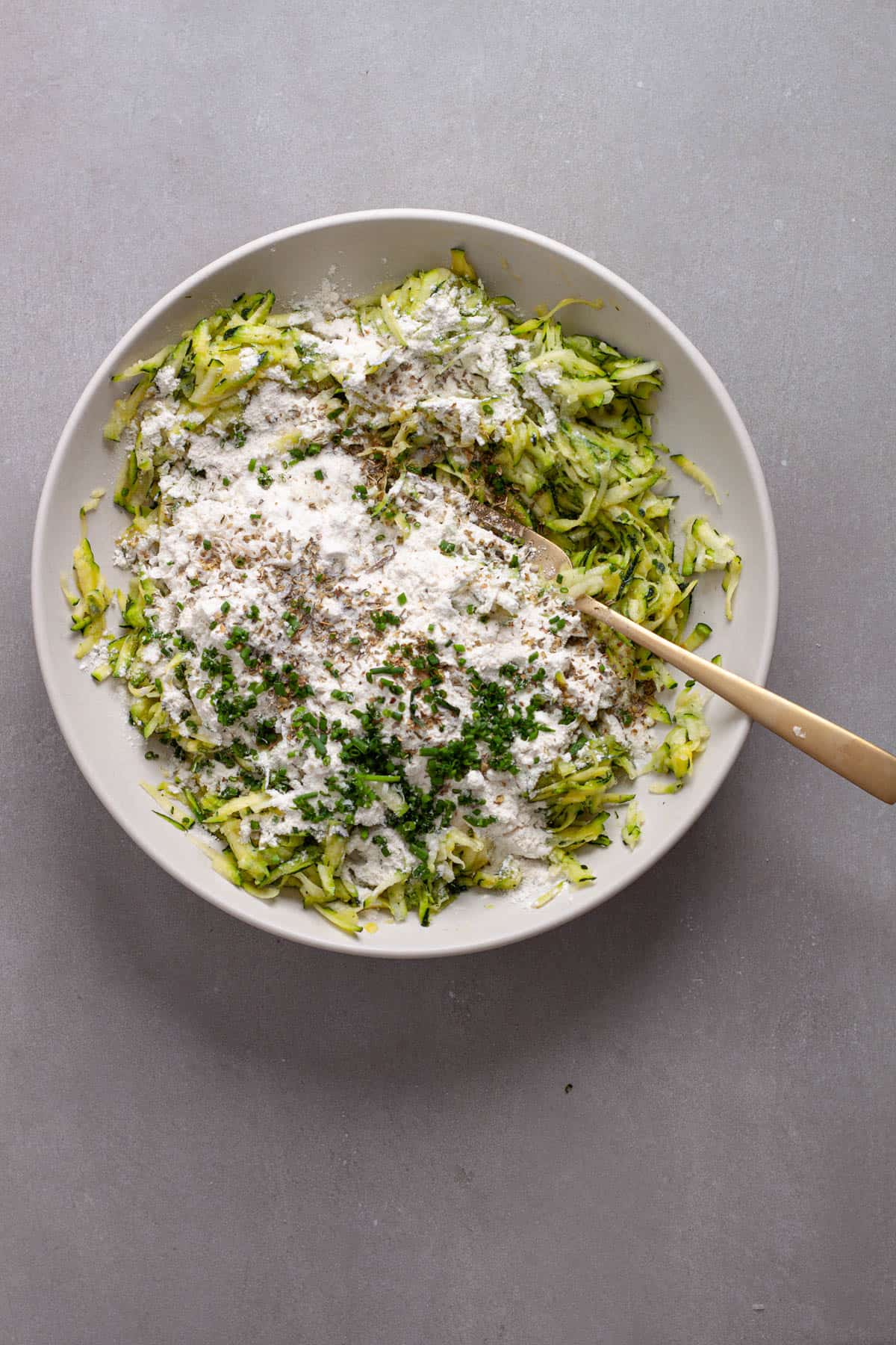 Shredded zucchini in a mixing bowl with eggs, flour and spices getting mixed in.