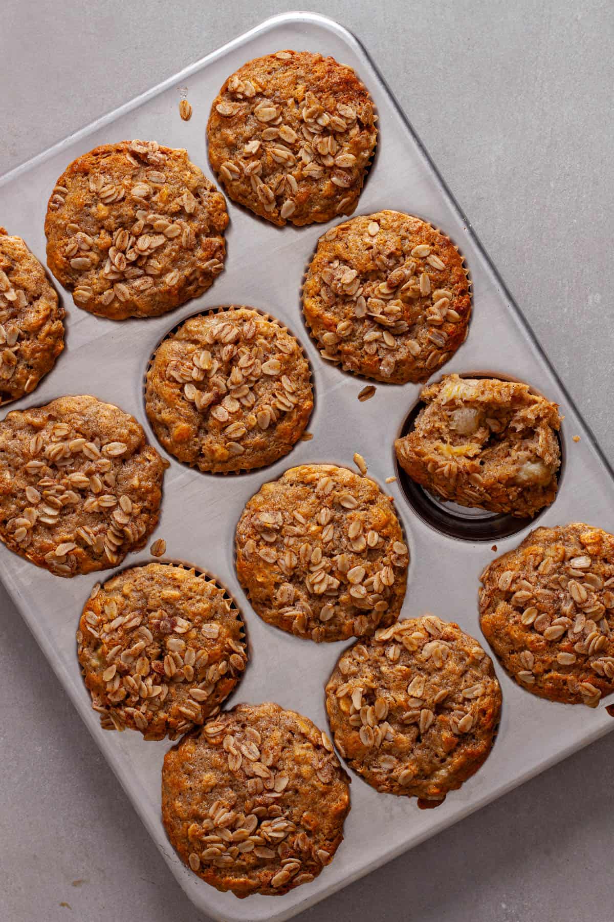A muffin tin of banana oat muffins with one on its side with a bite taken out.