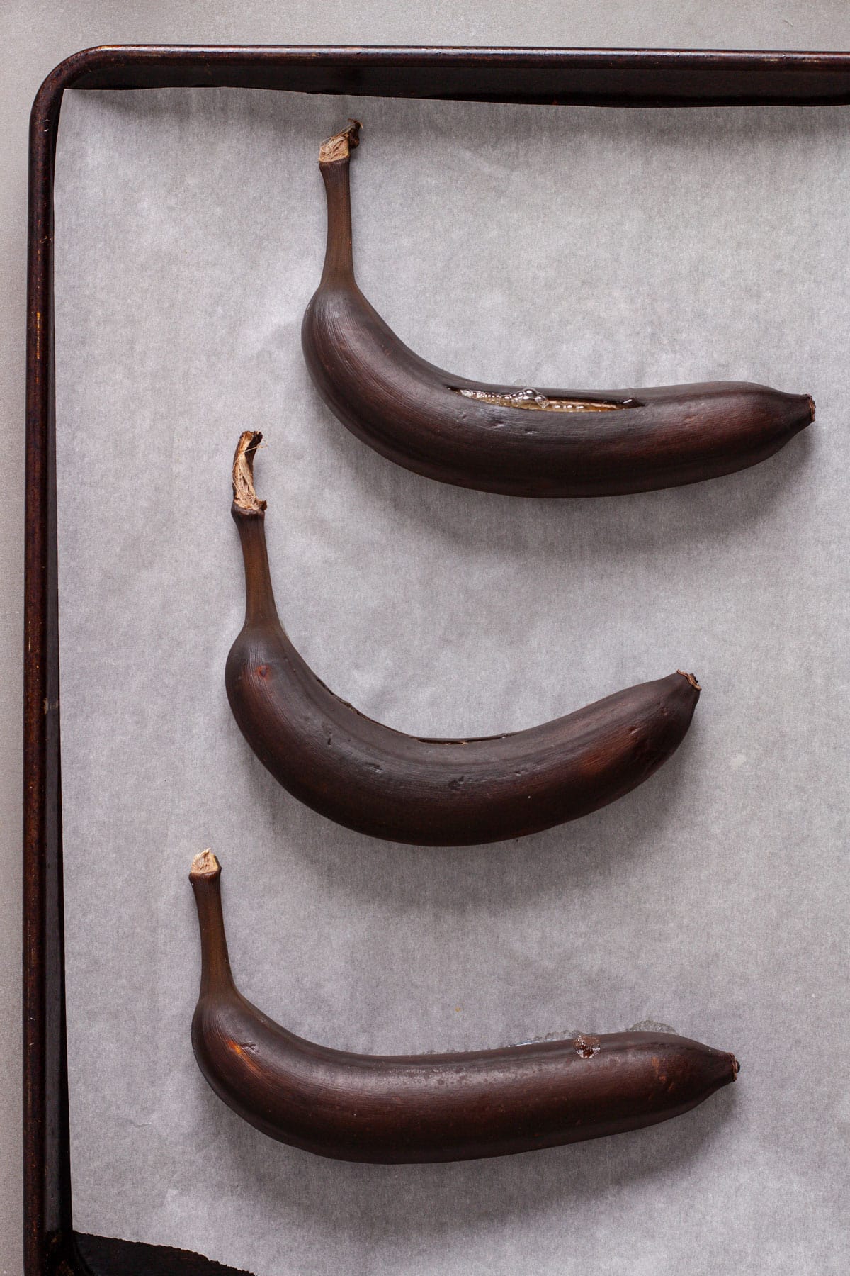 Blackened and roasted bananas in their peel on a parchment lined baking sheet.