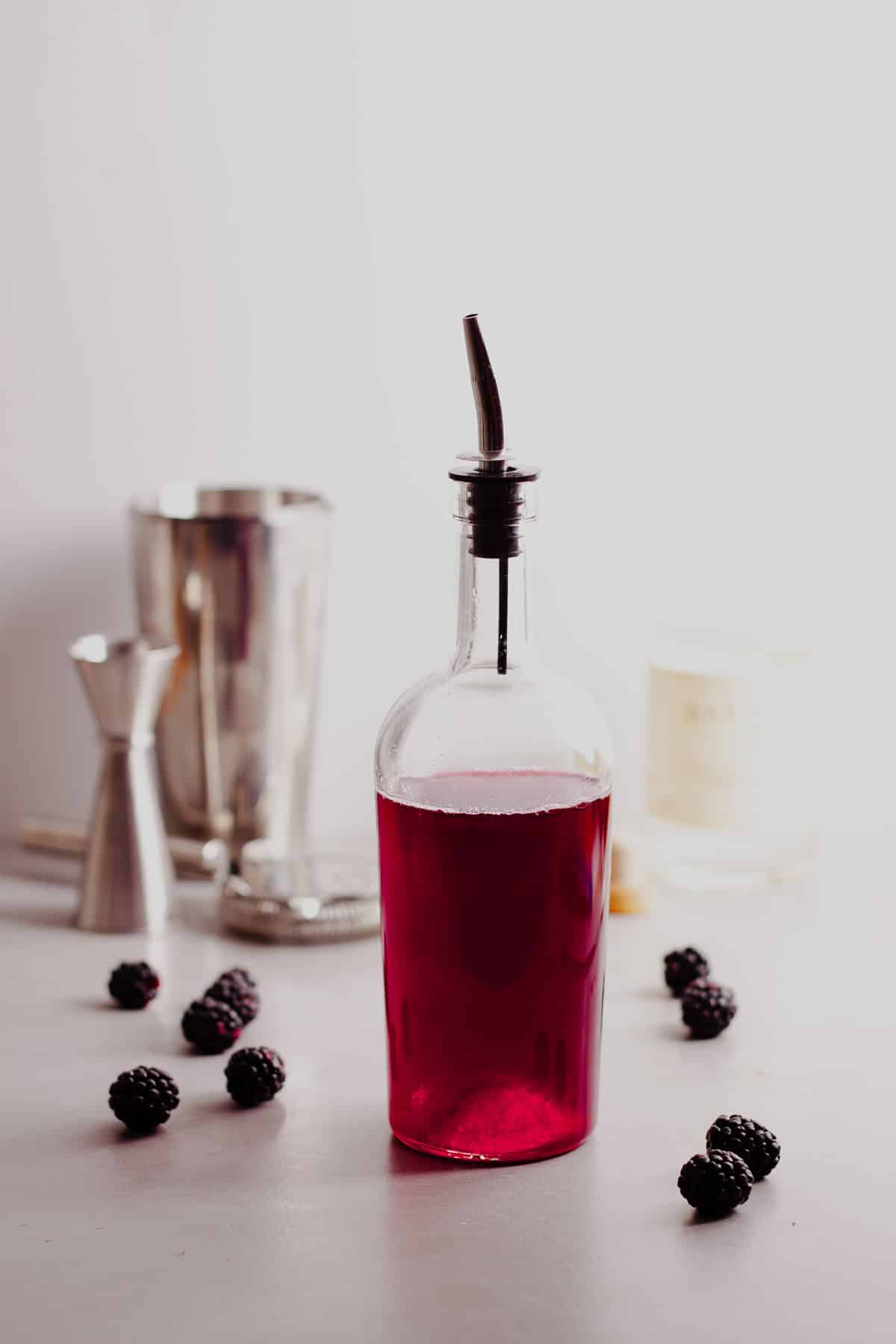 A glass bottle of blackberry simple syrup with blackberries on the side and cocktail instruments in the background.