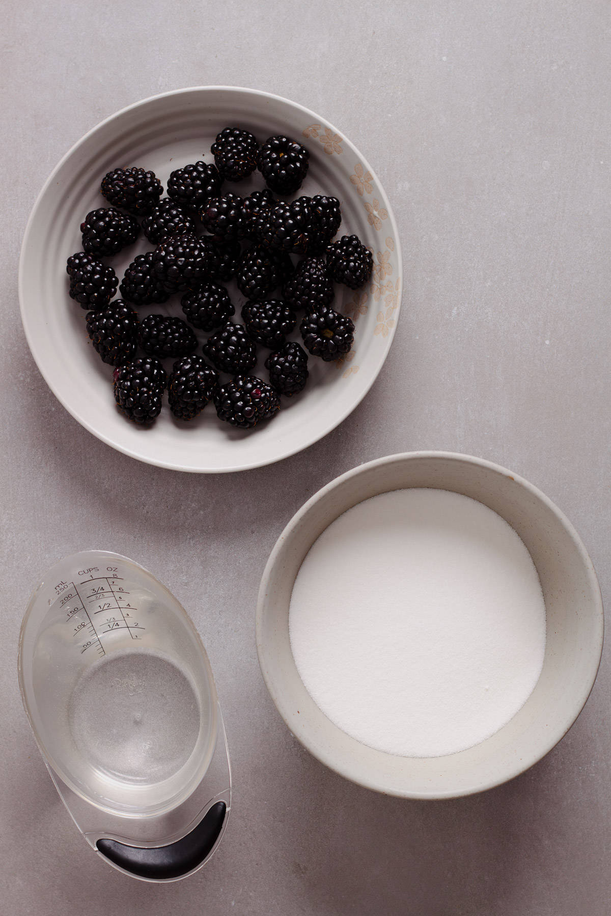 Fresh blackberries, sugar and water on a gray table for a blackberry simple syrup recipe.