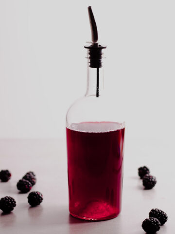 A glass bottle with a dark red blackberry simple syrup and fresh blackberries to the side.