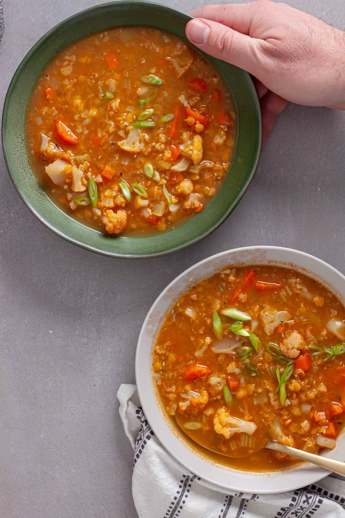 A couple bowls of cauliflower and lentil soup with a hand putting a bowl down on a gray table.