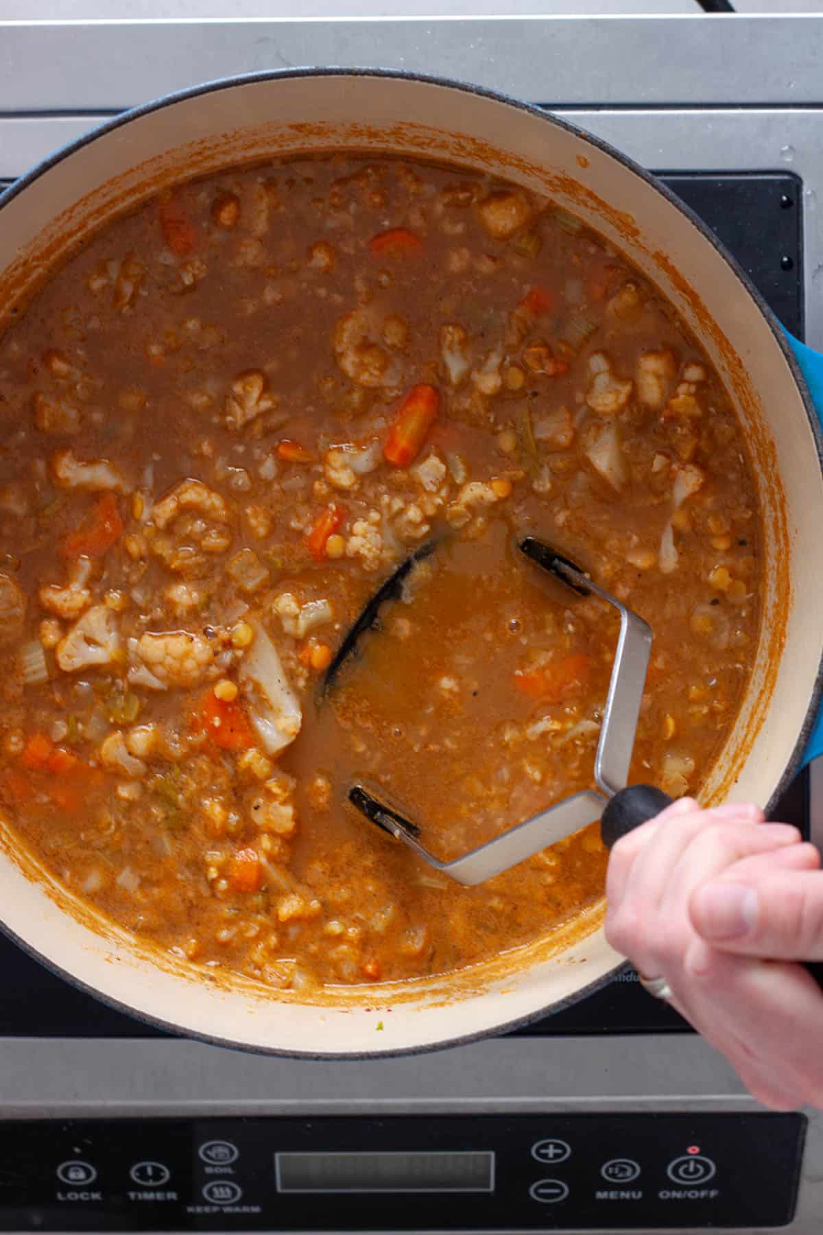 A potato masher gently smashing vegetables in a Dutch oven for a lentil soup with cauliflower.