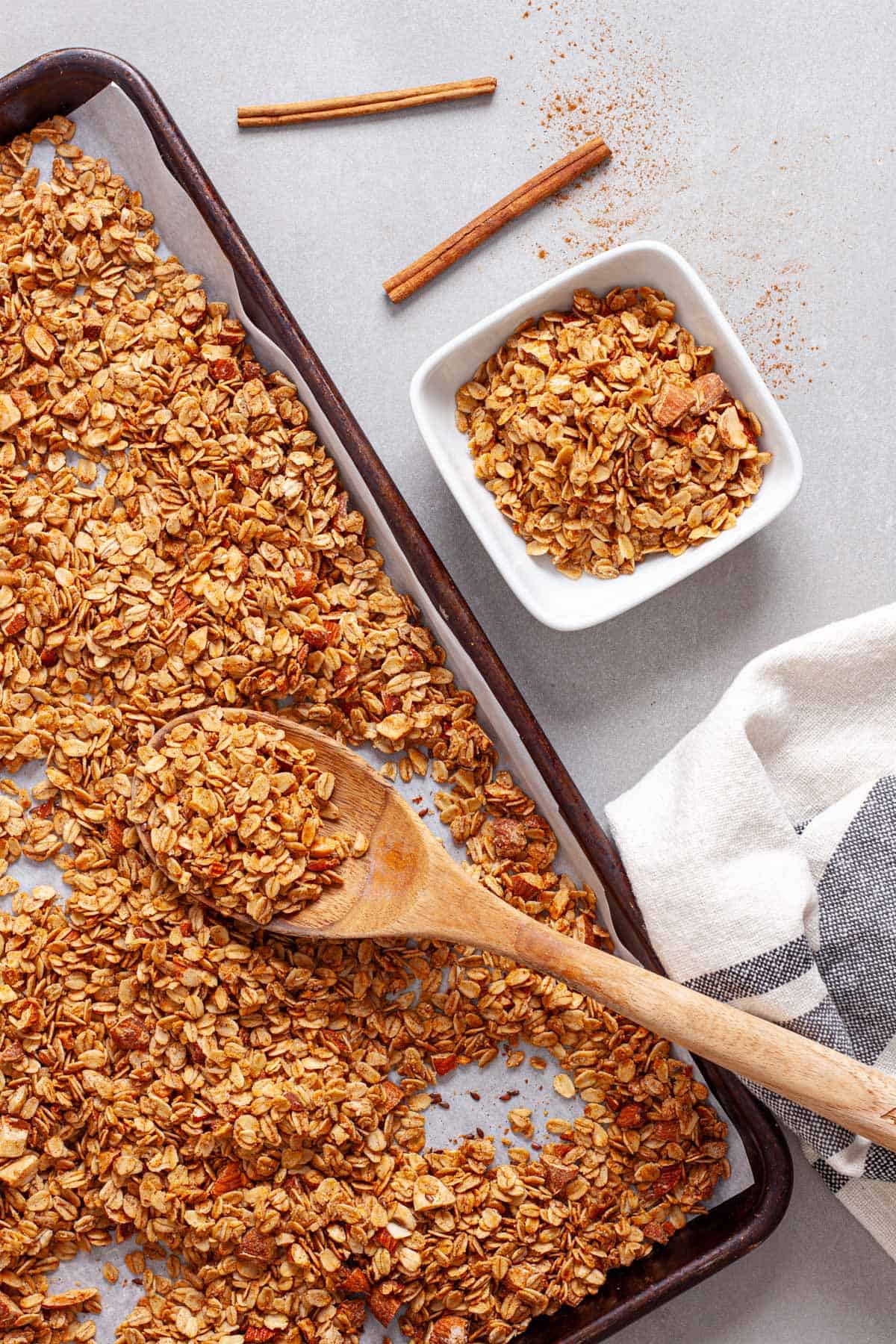 A wooden spoon on a sheet pan of homemade granola serving the granola into a small white bowl.