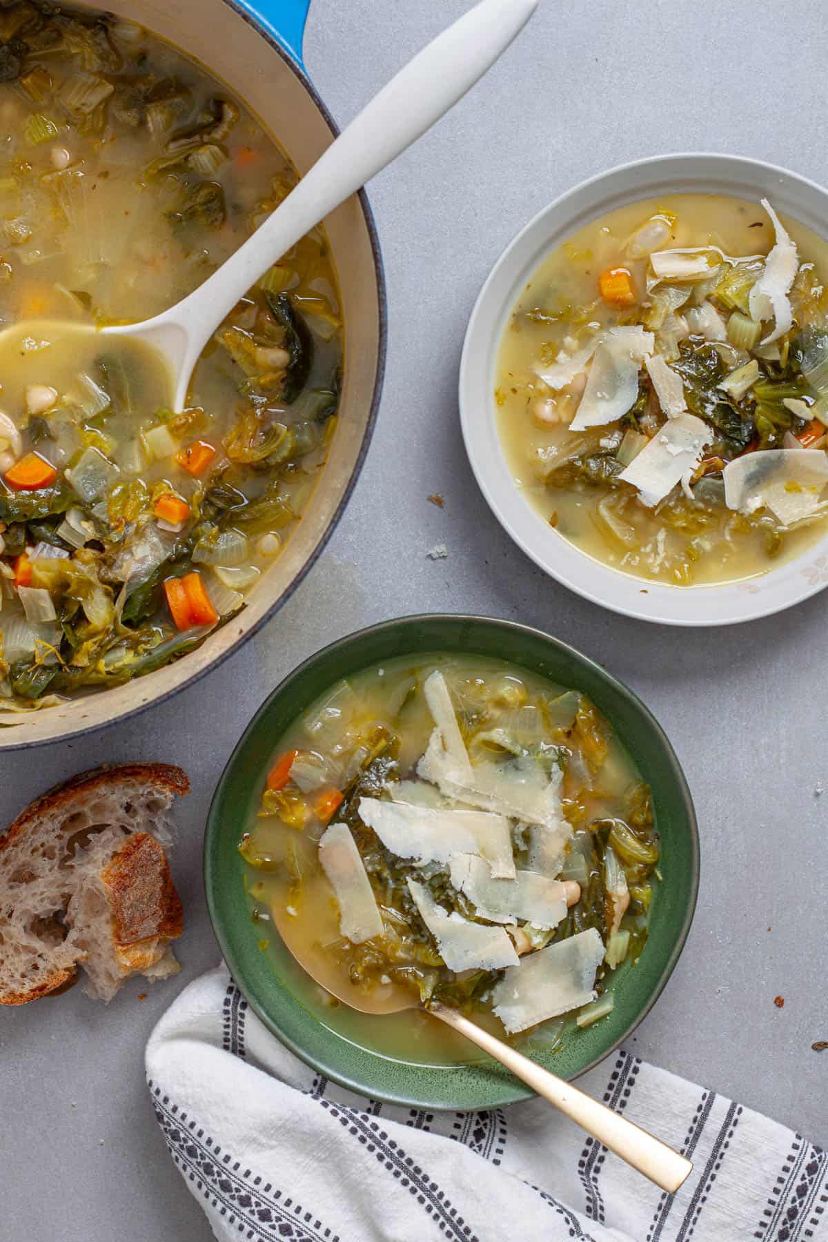 Two servings of escarole and white bean soup in small serving bowls with a pot of soup to the side.