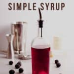 A glass bottle of blackberry simple syrup with blackberries on the side and cocktail instruments in the background.