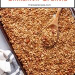 A baking sheet of homemade granola with a wooden spoon taking some out.