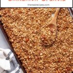 A baking sheet of homemade granola with a wooden spoon taking some out.