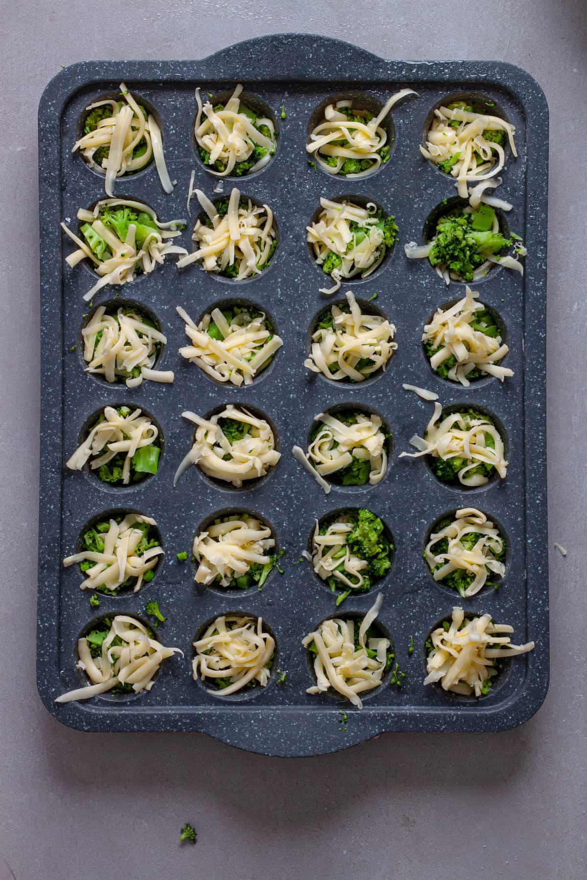 A mini muffin pan with shredded cheese and chopped broccoli.