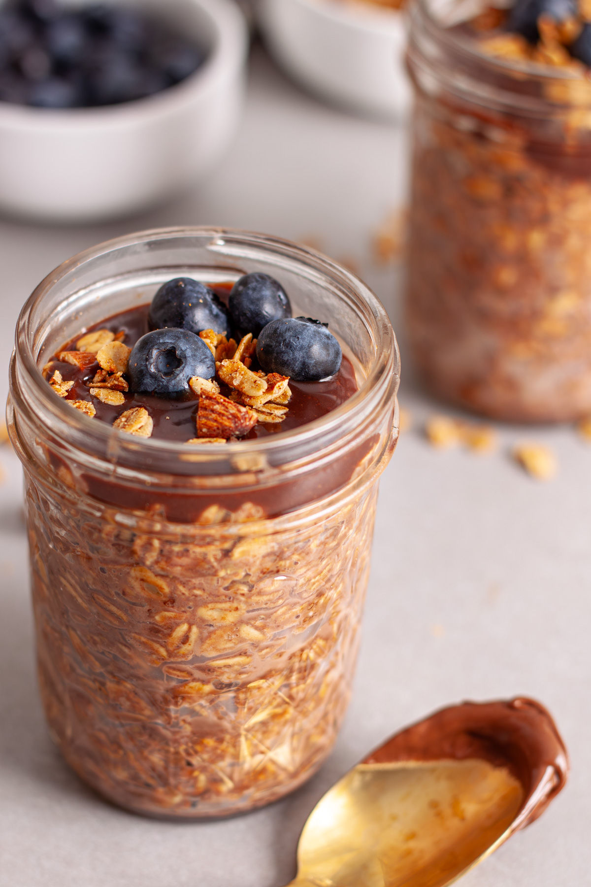 A close up of a small mason jar of Nutella overnight oats topped with blueberries and granola on a gray table.