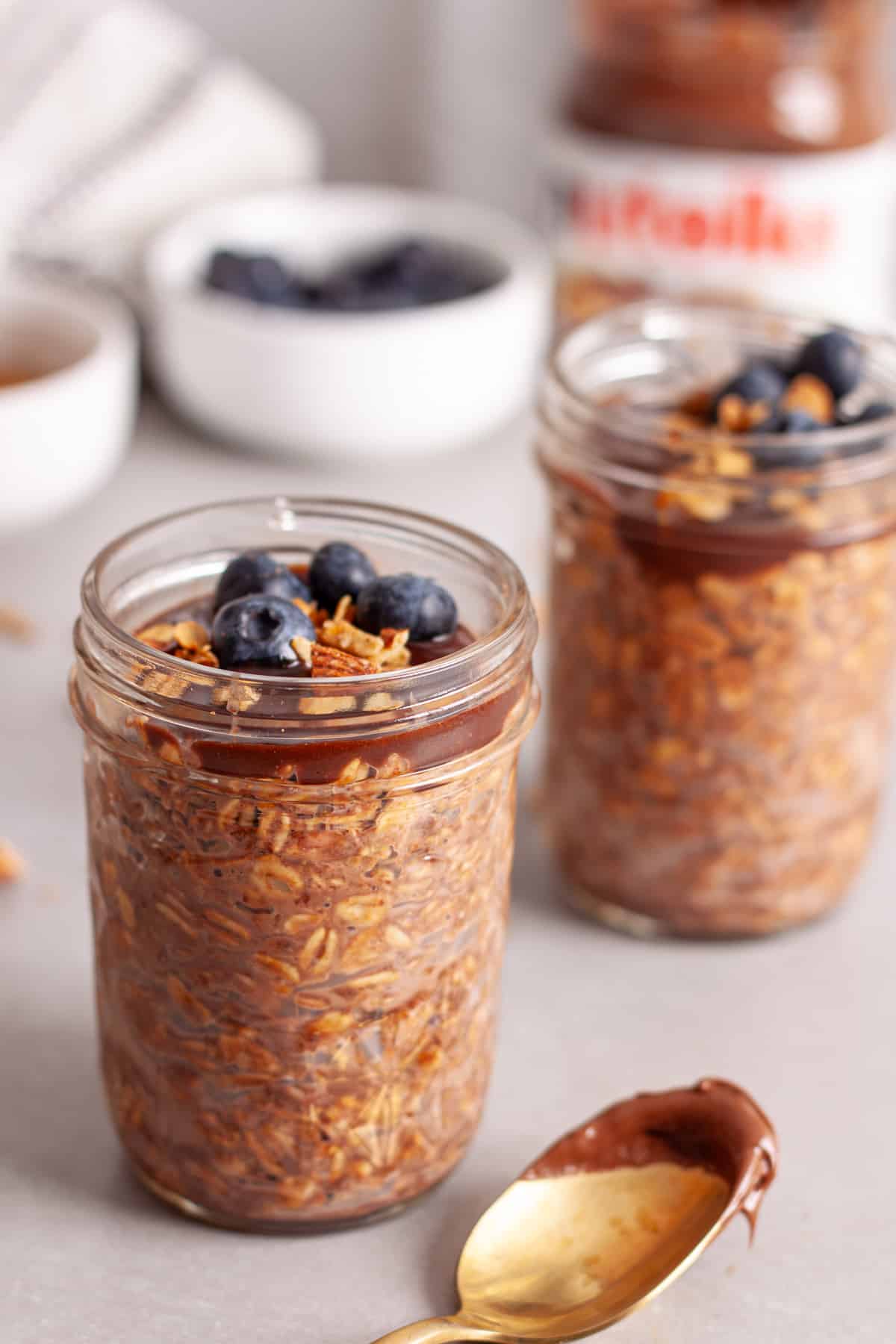 A couple mason jars with Nutella overnight oats topped with blueberries and granola on a gray table.
