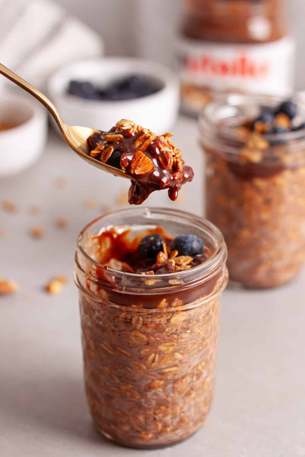 A spoon taking a bit of chocolate overnight oats out of a small mason jar on a gray table.