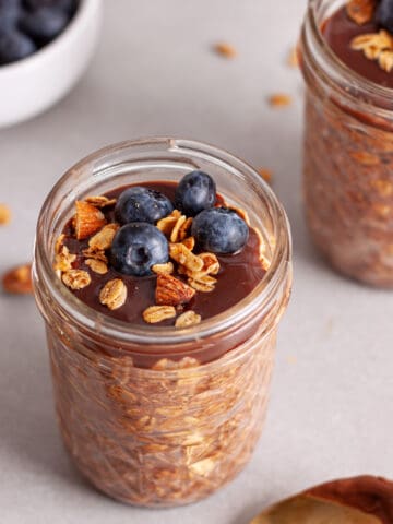 A single mason jar portion of Nutella overnight oats topped with blueberries and granola.