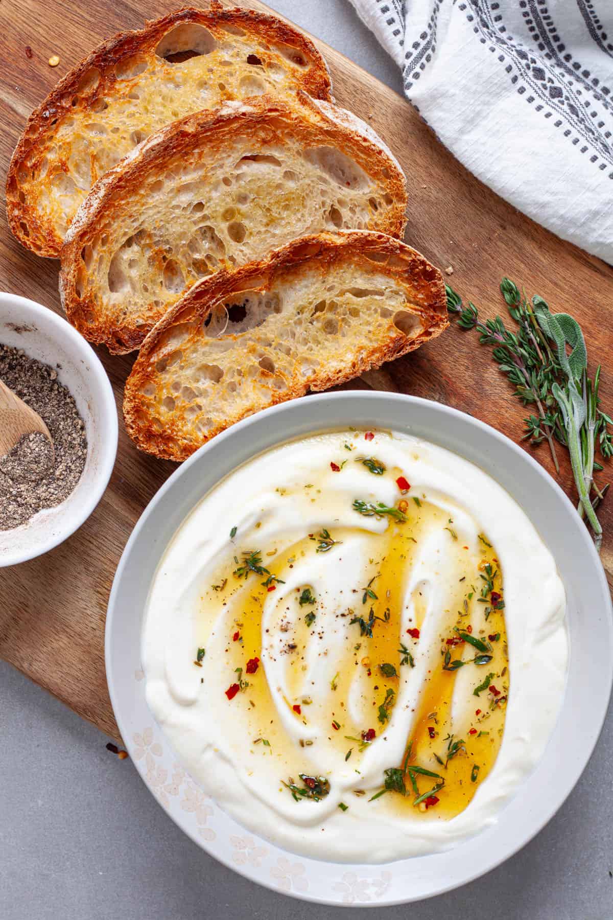 A bowl of whipped ricotta topped with honey and herbs on a cutting board with toasted slices of bread.