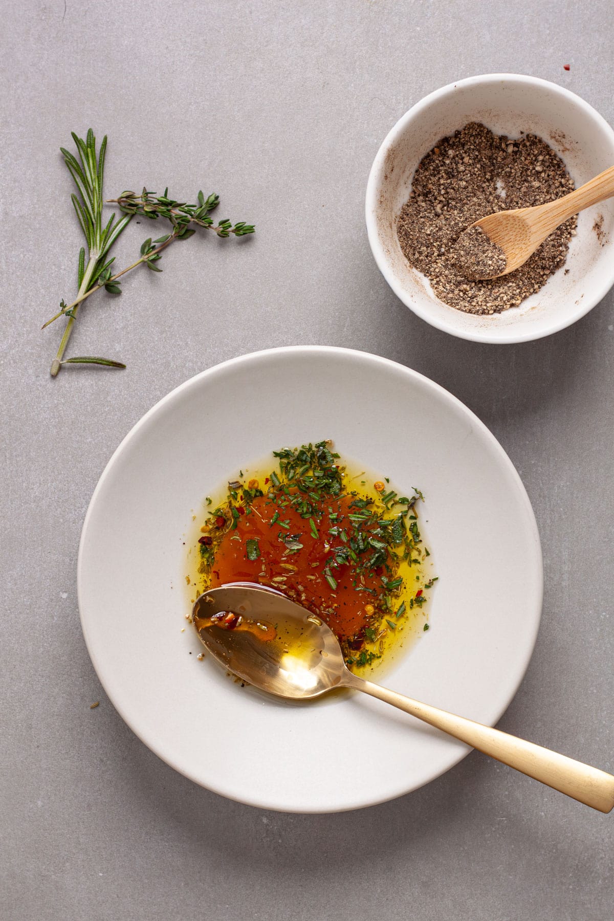 A small bowl with honey, oil, herbs and crushed red pepper flakes on a gray table.