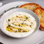 A serving bowl of whipped ricotta dip topped with honey and herbs on a plate with toasted bread.