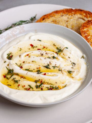 A serving bowl of whipped ricotta dip topped with honey and herbs on a plate with toasted bread.