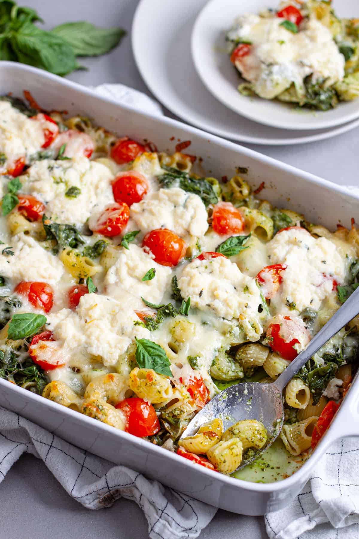 A spoon taking a serving of ricotta pesto pasta out of a white casserole dish.