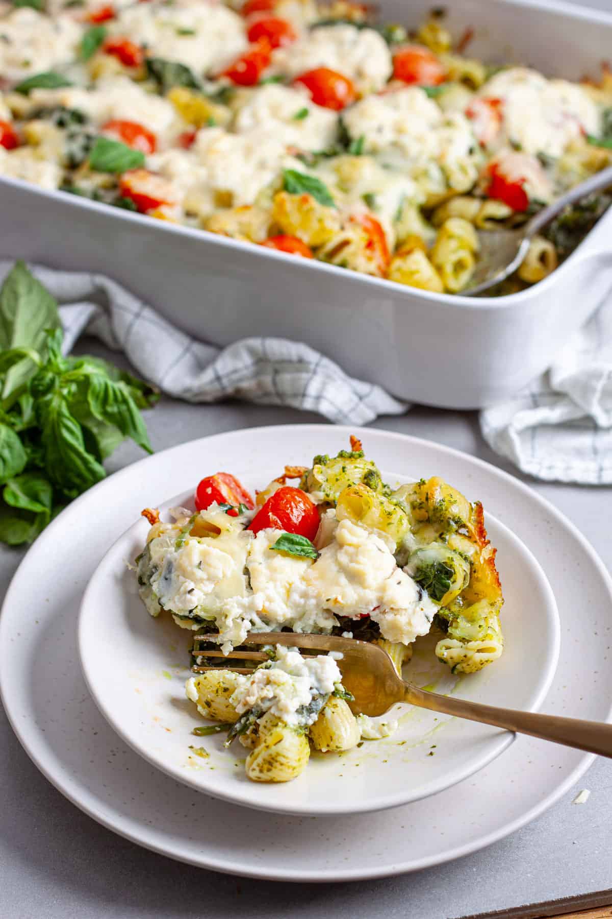 A serving of ricotta pesto pasta on a plate with a casserole dish of the baked pasta in the background.