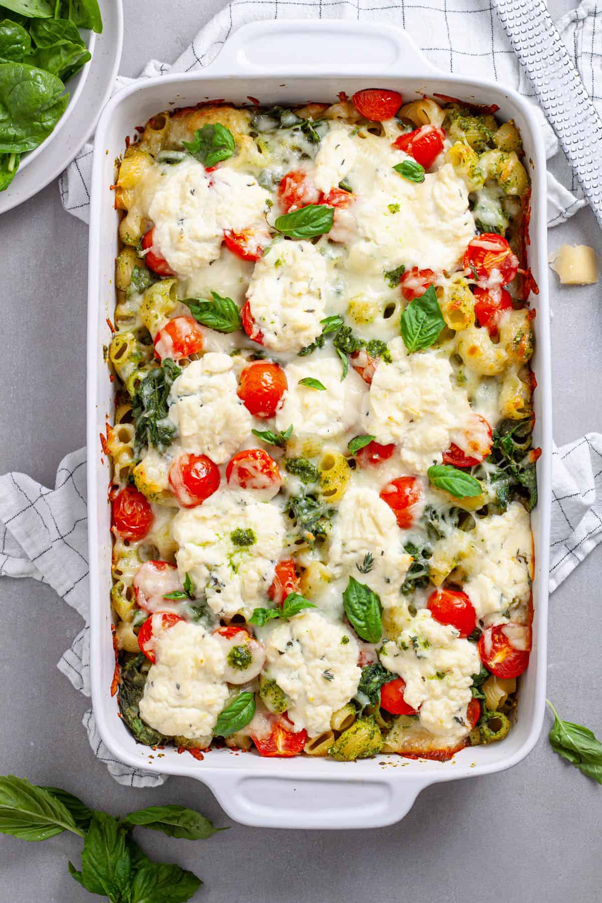 A casserole dish of ricotta pesto pasta with tomatoes and spinach on a gray table.