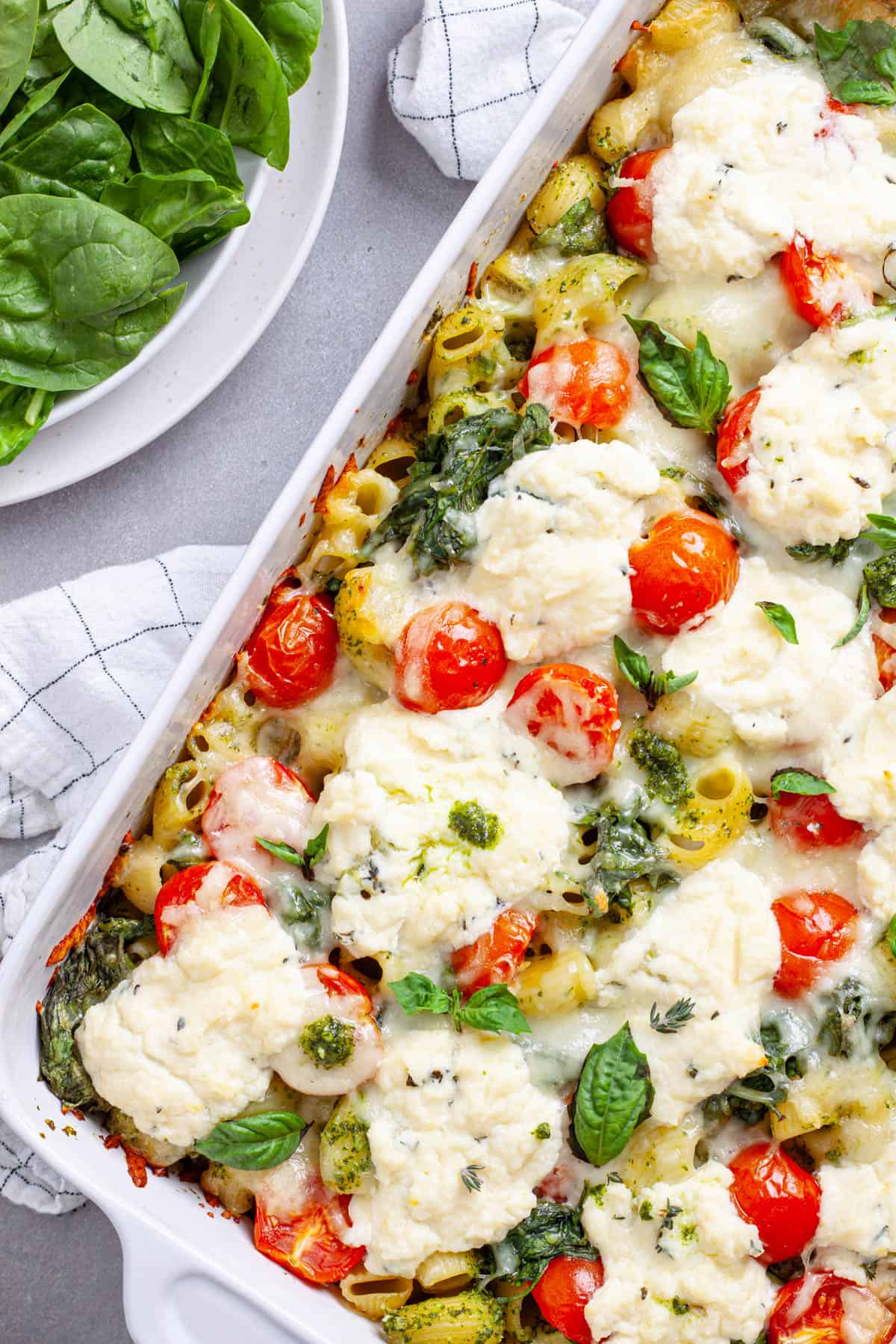 A white casserole dish with ricotta pesto pasta topped with mozzarella, tomatoes and spinach.