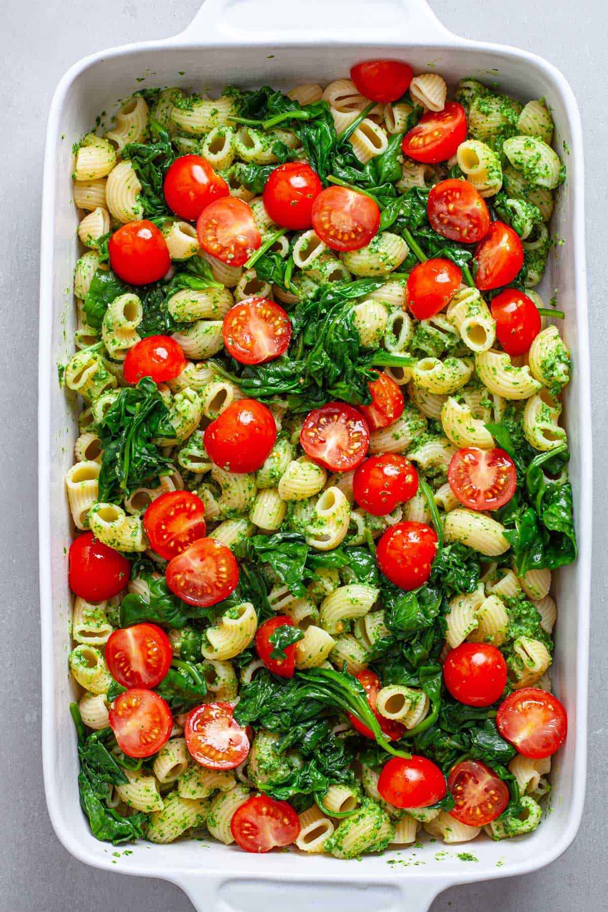 Cherry tomatoes and wilted spinach on top of a casserole dish of pasta and pesto.