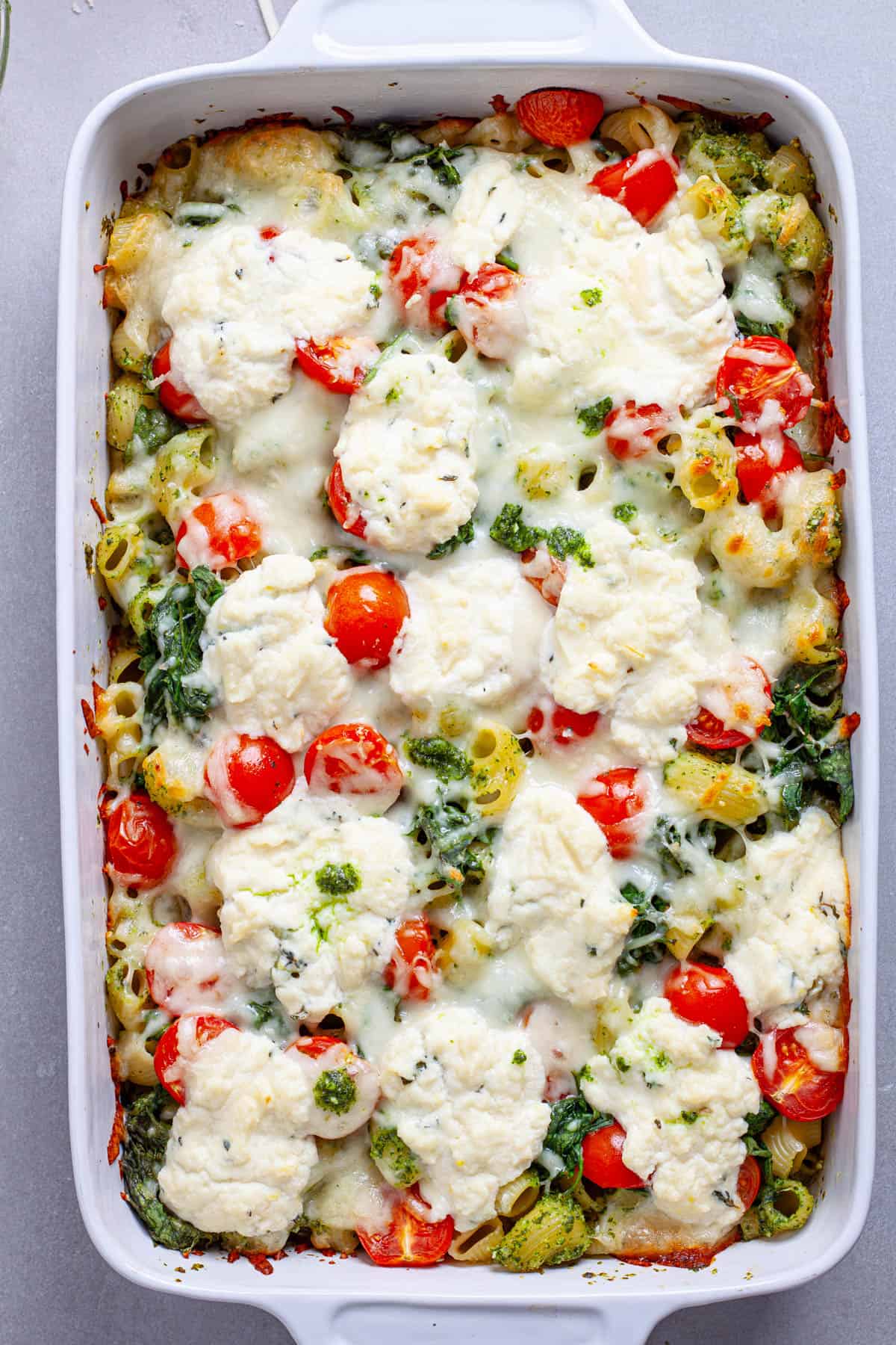 A casserole dish of pesto and ricotta baked pasta hot out of the oven topped with tomatoes and spinach.