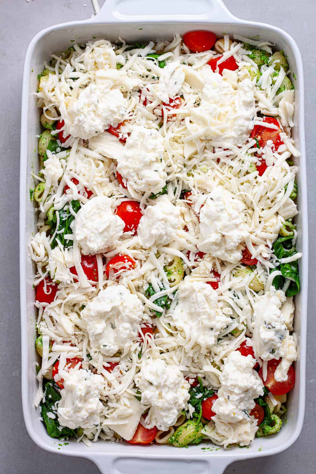 A casserole dish of pasta and pesto with ricotta and mozzarella scattered on top, along with tomatoes and spinach.