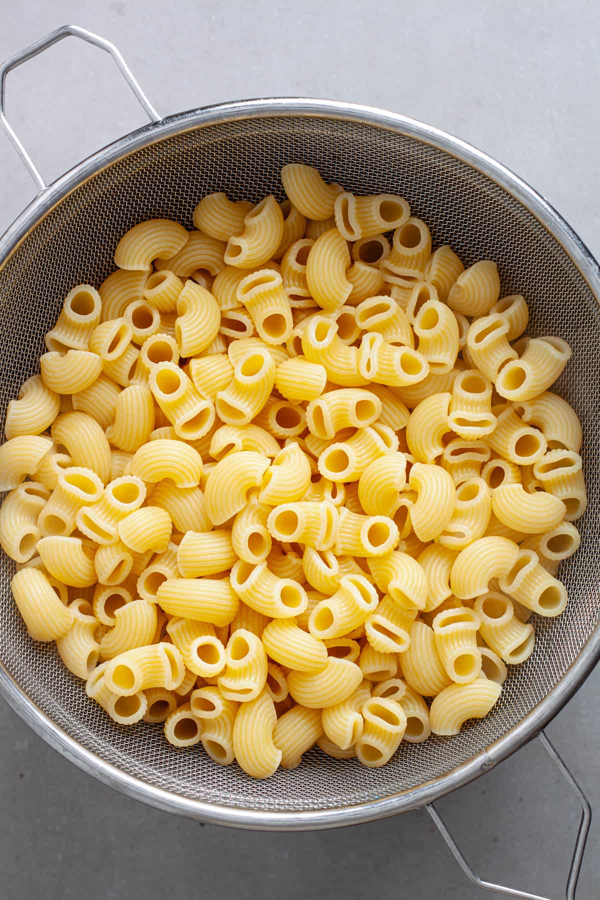 Cooked pasta cooling in a strainer on a gray table.