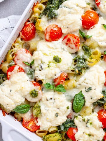 A close up of a baked ricotta pesto pasta topped with tomatoes, spinach and melty cheese.