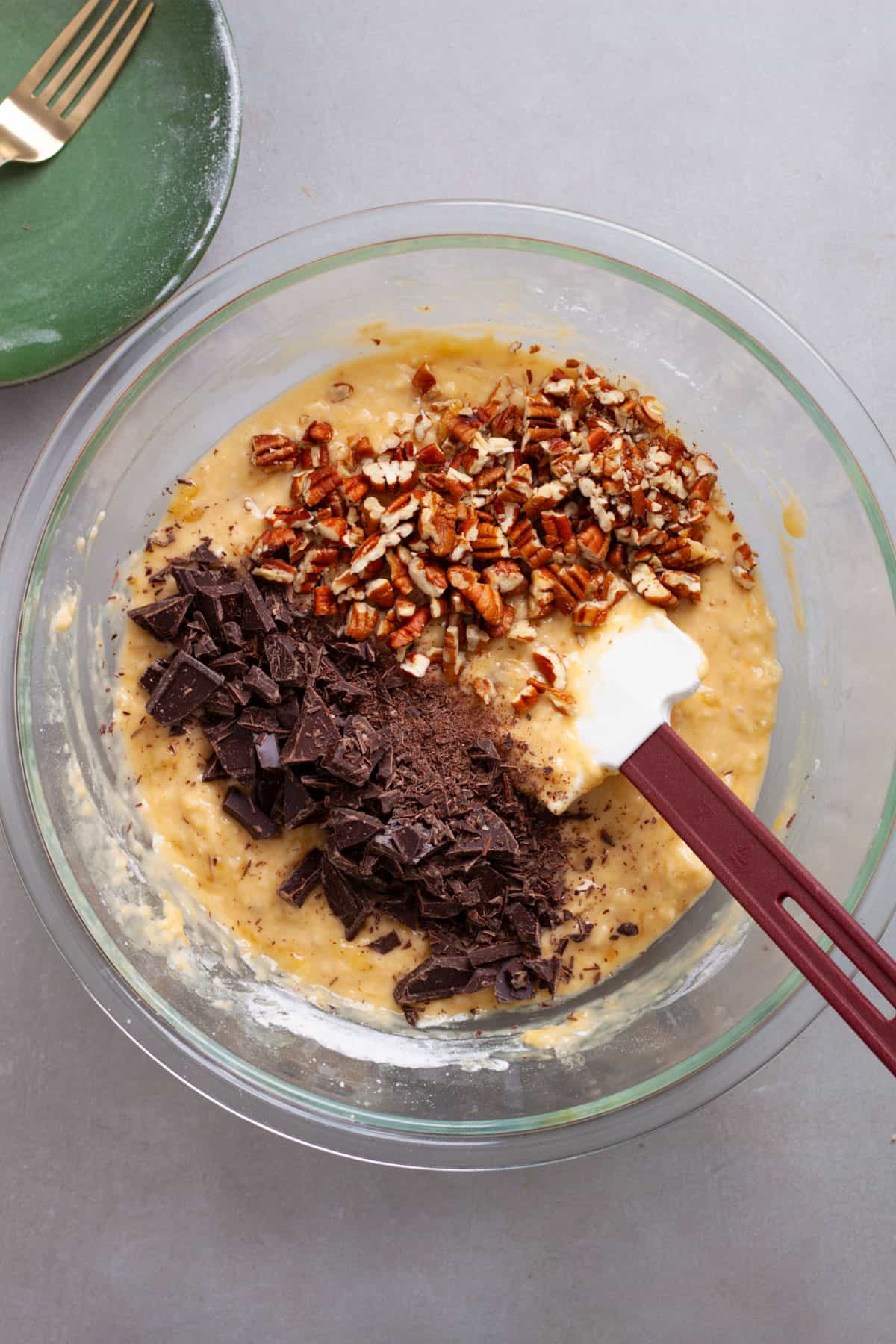 Chocolate chunks and chopped pecans added to banana muffin batter in a glass bowl.