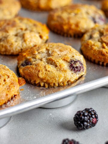 Banana blackberry oatmeal muffins in a muffin tin with loose fresh blackberries in front.
