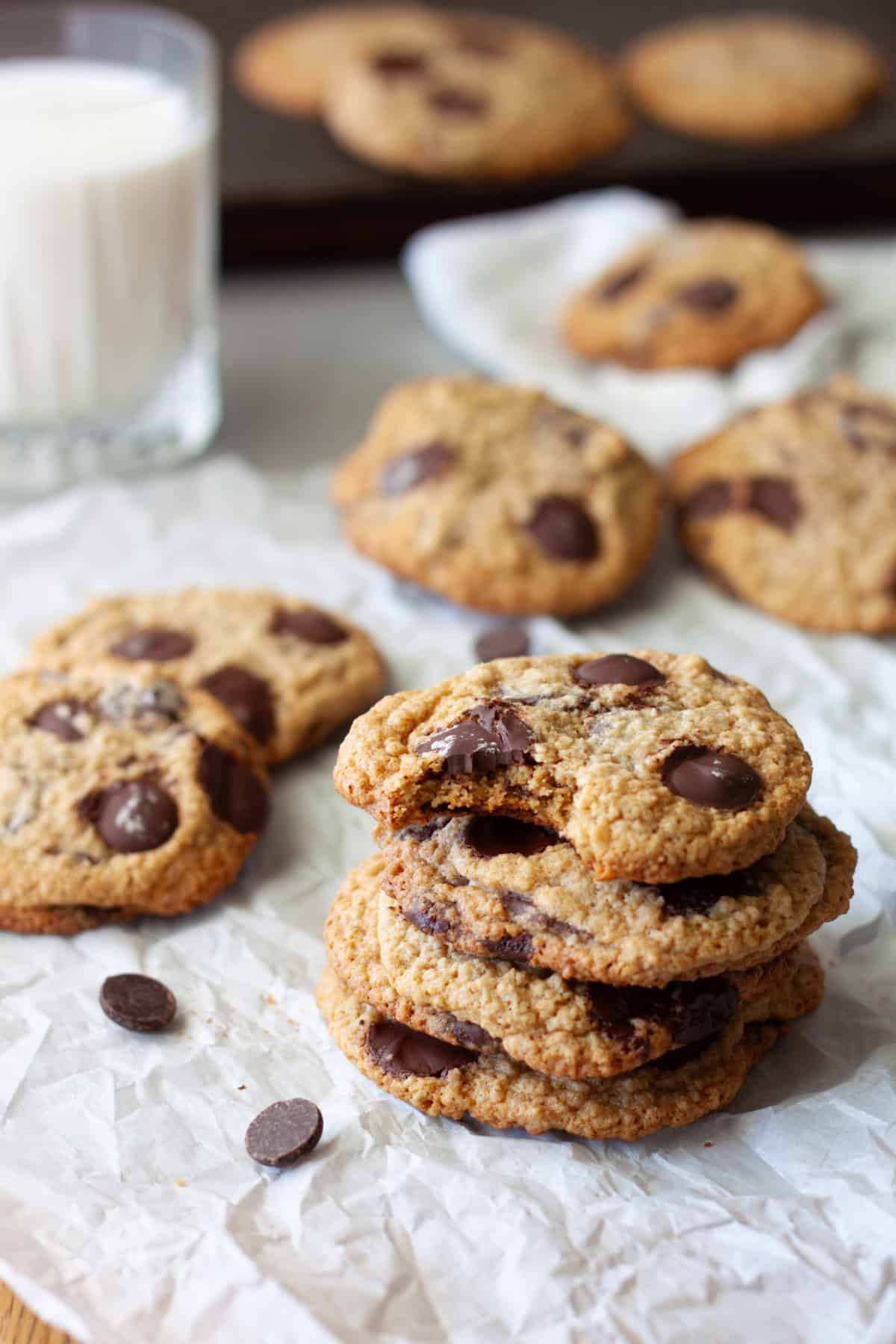 A stack of oat flour chocolate chip cookies on parchment paper with more in the background and a glass of milk to the side.